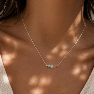 May include: A delicate silver chain necklace featuring three small, round beads centered on the strand. The beads consist of a pale green stone, a translucent white opal-like stone, and a warm yellow citrine-colored stone, separated by tiny silver spacers.