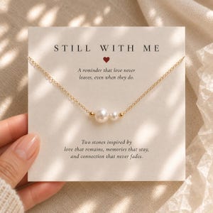 Può includere: Una collana a catena dorata con due perle bianche di dimensioni diverse, presentata su un cartoncino bianco con il testo STILL WITH ME, un cuore rosso e un messaggio sull'amore. La collana presenta piccole perline dorate accanto alle perle.
