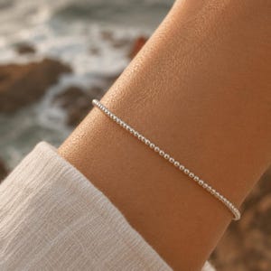Pode incluir: Uma delicada pulseira de contas prateadas. A pulseira é composta por pequenas contas redondas e espaçadas. A pulseira é usada em um pulso, com um tecido branco macio ao fundo. O oceano é visível ao fundo.