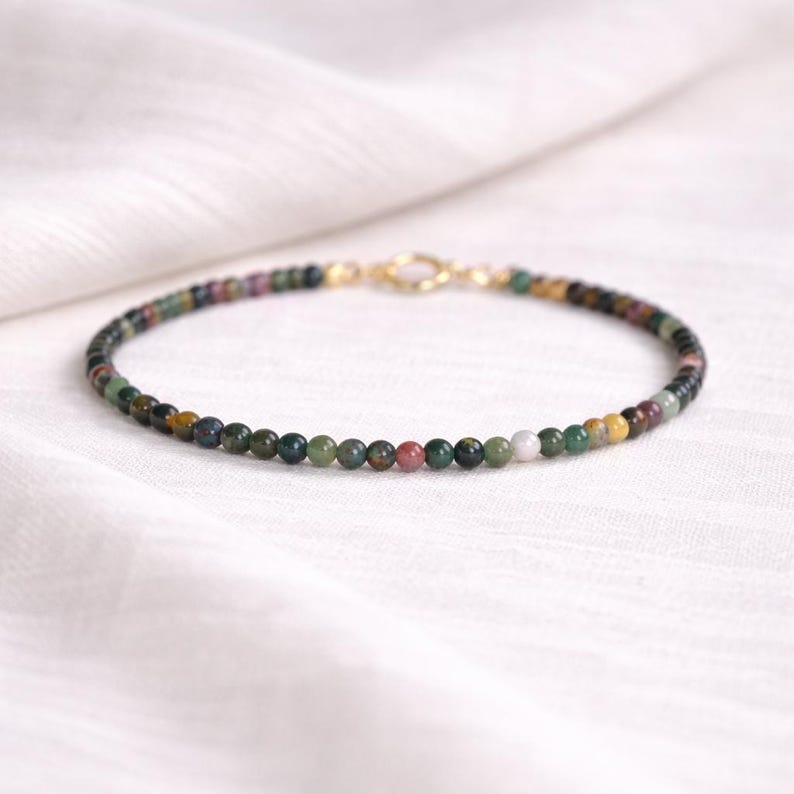 May include: A delicate bracelet composed of small, round, multi-colored gemstone beads in shades of green, brown, red, and yellow. The bracelet features a gold-toned circular spring ring clasp that connects the ends of the beaded strand.