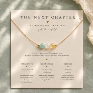 Op de afbeelding: Gouden ketting met drie edelstenen: lichtgroen, wit en geel. De kaart toont de tekst "THE NEXT CHAPTER" en "yet to unfold". Gepresenteerd op een kaart.