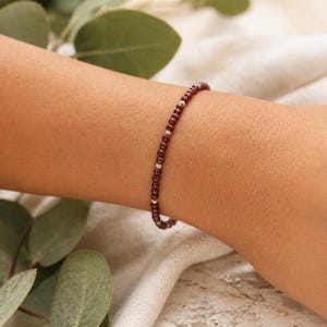 Pode incluir: Uma pulseira delicada com pequenas contas redondas de cor vermelho escuro e contas prateadas. A pulseira é usada no pulso, mostrando seu design simples. O fundo apresenta tons suaves e neutros e vegetação.