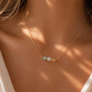 May include: A delicate gold chain necklace featuring three small, smooth, round gemstone beads centered on the strand. The beads consist of a pale green stone, a translucent white stone, and a warm yellow stone, all held by gold metal accents.