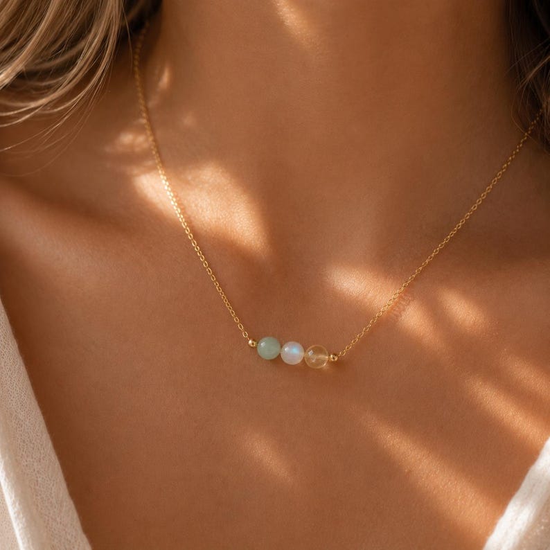 May include: A delicate gold chain necklace featuring three small, smooth, round gemstone beads centered on the strand. The beads consist of a pale green stone, a translucent white stone, and a warm yellow stone, all held by gold metal accents.