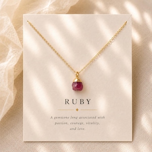 May include: A gold chain necklace featuring a raw, deep red ruby gemstone pendant with a gold cap. The necklace is displayed on a cream-colored card with the text RUBY and A gemstone long associated with passion, courage, vitality, and love.