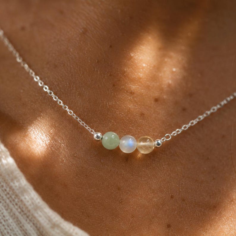 May include: A delicate silver chain necklace featuring three small, round, polished gemstone beads in a row. The beads consist of a pale green stone, a translucent white moonstone with a blue sheen, and a warm yellow citrine stone.