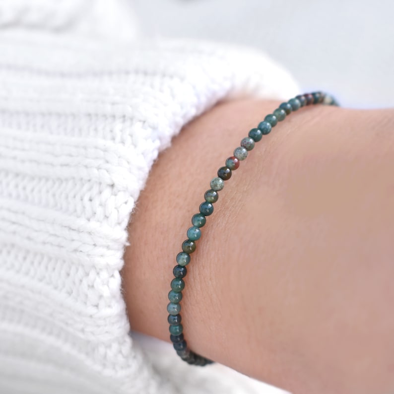 May include: A delicate bracelet composed of small, round, multi-colored gemstone beads in shades of deep forest green, teal, and subtle hints of earthy red. The beads are strung together in a single strand, creating a minimalist and refined accessory.