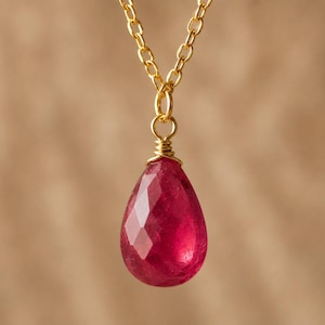 May include: A gold-colored chain necklace with a faceted, teardrop-shaped, red gemstone pendant. The pendant is wire-wrapped and hangs from a small gold loop. The background is a solid, warm brown.