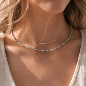 May include: A two-strand necklace set. The top necklace is a delicate gold chain. The bottom necklace features small, light blue-green beads. The necklaces are worn around the neck.