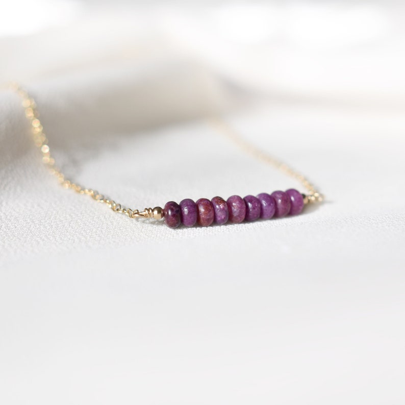 May include: A delicate gold chain necklace featuring a row of small, round, deep purple gemstones. The gemstones are evenly spaced and create a linear design. The necklace is displayed on a white, textured surface.