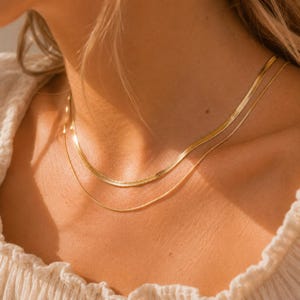 May include: A layered gold necklace set consisting of a flat, wide herringbone chain and a thin, delicate cable chain. Both chains feature a polished, reflective finish that catches the light, resting against the collarbone in a tiered arrangement.