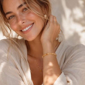 May include: A smiling person wearing a gold necklace and a beaded bracelet. The bracelet and necklace feature small, round, amber-colored beads. The person is wearing a light-colored top and has blonde hair.