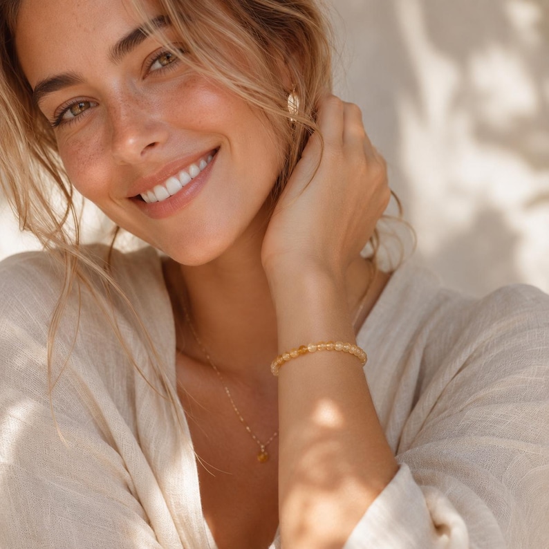May include: A smiling person wearing a gold necklace and a beaded bracelet. The bracelet and necklace feature small, round, amber-colored beads. The person is wearing a light-colored top and has blonde hair.