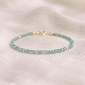 May include: A delicate bracelet with small, round, pale blue beads and gold-coloured accents. The bracelet has a gold clasp and is displayed on a soft, off-white fabric. This piece of jewellery is a stylish accessory.