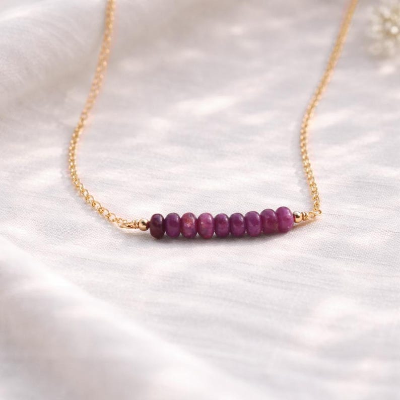 May include: A delicate gold chain necklace featuring a row of small, oval-shaped, deep red gemstones. The gemstones are evenly spaced and set horizontally. The necklace is displayed on a white, textured surface.