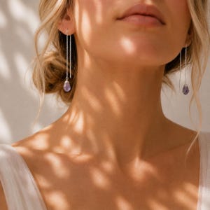 May include: A pair of silver threader earrings featuring delicate chains that pass through the earlobe, each ending in a small, raw-cut, light purple amethyst gemstone pendant with a metallic silver cap, creating a long, dangling silhouette.
