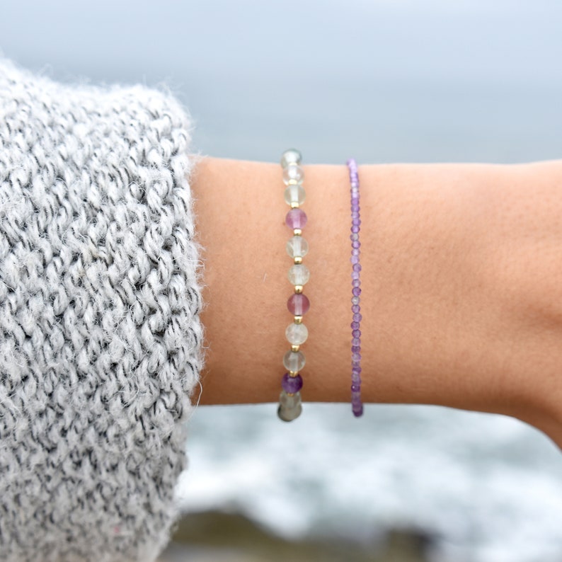 May include: Two beaded bracelets on a wrist. One bracelet features a mix of clear, purple, and gold beads. The other is a delicate strand of small purple beads. The background is a blurred view of the ocean.