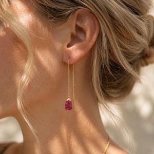 May include: A gold threader earring featuring a delicate fine chain that passes through the earlobe, ending in a raw, deep pink gemstone pendant with a textured, irregular shape and a small gold cap at the top of the stone.