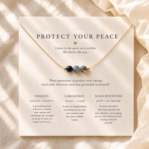 May include: A gold chain necklace featuring three small round beads: one black obsidian, one iridescent labradorite, and one grey black moonstone. The necklace is displayed on a white card with the text PROTECT YOUR PEACE and descriptions of the stones.