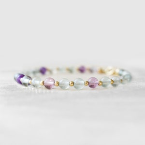 May include: A beaded bracelet featuring a mix of colorful fluorite stones in shades of purple, pink, and green. The bracelet is strung with small gold-colored beads. The bracelet is a delicate piece of jewelry.