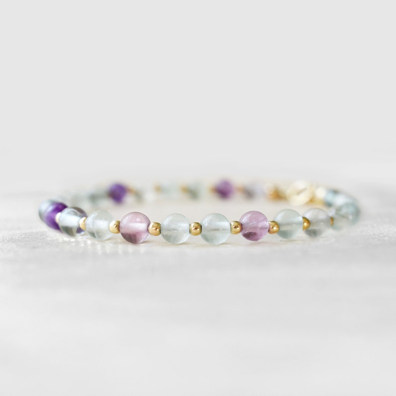 May include: A beaded bracelet featuring a mix of colorful fluorite stones in shades of purple, pink, and green. The bracelet is strung with small gold-colored beads. The bracelet is a delicate piece of jewelry.