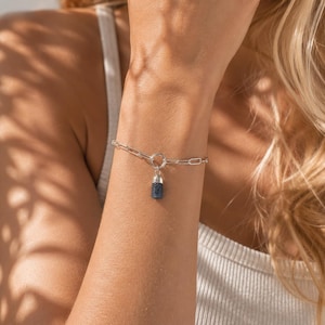 Peut inclure: Un bracelet en chaîne argentée composé de maillons ovales allongés et d'un anneau circulaire central. Un petit pendentif cylindrique en pierre bleu foncé avec un capuchon argenté est suspendu à l'anneau central, formant un bijou raffiné.