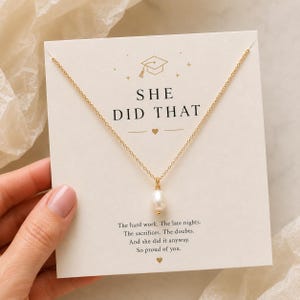 May include: A gold-colored necklace with a single pearl pendant is displayed on a card. The card has the text "SHE DID THAT" and a graduation cap graphic. The card also includes a motivational message.