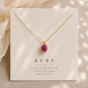 May include: A gold chain necklace featuring a small, raw, deep pink ruby gemstone pendant. The necklace is displayed on a white card with the text RUBY and a description of the gemstone's associations printed in black ink below the pendant.