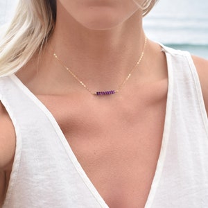 May include: A delicate gold necklace featuring a row of small, vibrant purple gemstones. The necklace is displayed on a person wearing a white top, with a blurred ocean background. The gemstones are evenly spaced along the gold chain.