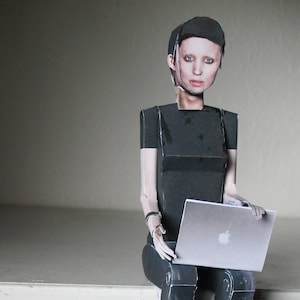 Può includere: Una figura di carta di una persona seduta con un computer portatile. La figura indossa una camicia nera e pantaloni neri. Il computer portatile è argentato e ha un logo Apple bianco.