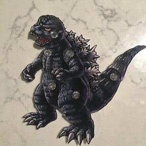 May include: A black and gray papercraft model of Godzilla, a fictional monster, with a textured surface and silver rivets.