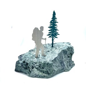 May include: A silver-toned metal silhouette of a hiker with a backpack and trekking poles standing next to a dark green metal pine tree. Both figures are mounted on a large, textured, natural green stone base with a rough, uneven surface.