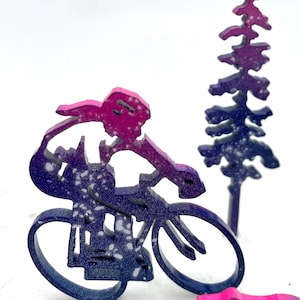 以下が含まれることがあります： 背景に松の木のある、自転車に乗っている人の木製のシルエット。シルエットはピンクとパープルの色合いで、白い斑点がある。