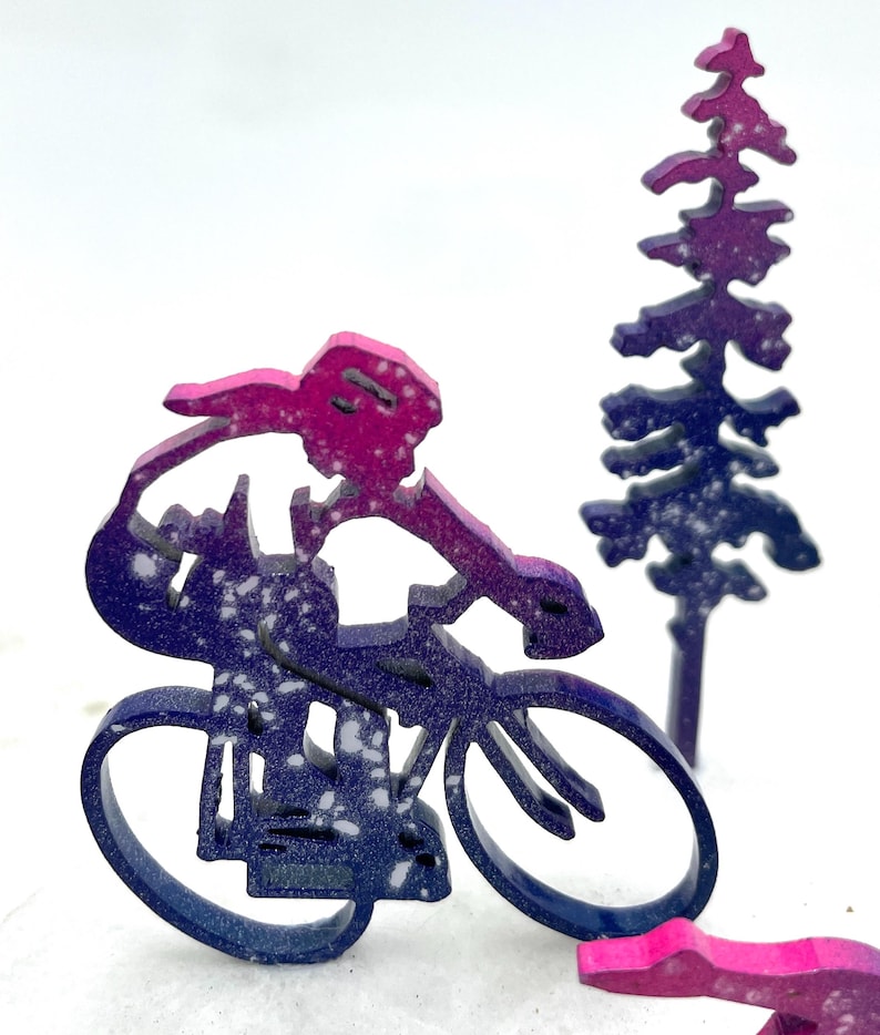 以下が含まれることがあります： 背景に松の木のある、自転車に乗っている人の木製のシルエット。シルエットはピンクとパープルの色合いで、白い斑点がある。