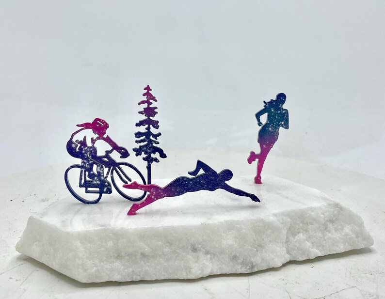 以下が含まれることがあります： 白い大理石の台座に、自転車に乗った人、泳いでいる人、走っている人の3つの金属製のフィギュアが置かれています。フィギュアはすべてシルエットになっており、ピンクとブルーのグラデーションになっています。