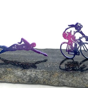 以下が含まれることがあります： 濃い灰色の石の表面に、水泳選手と自転車に乗った人の金属製のシルエットが2つあります。シルエットはピンクとパープルのグラデーションになっています。