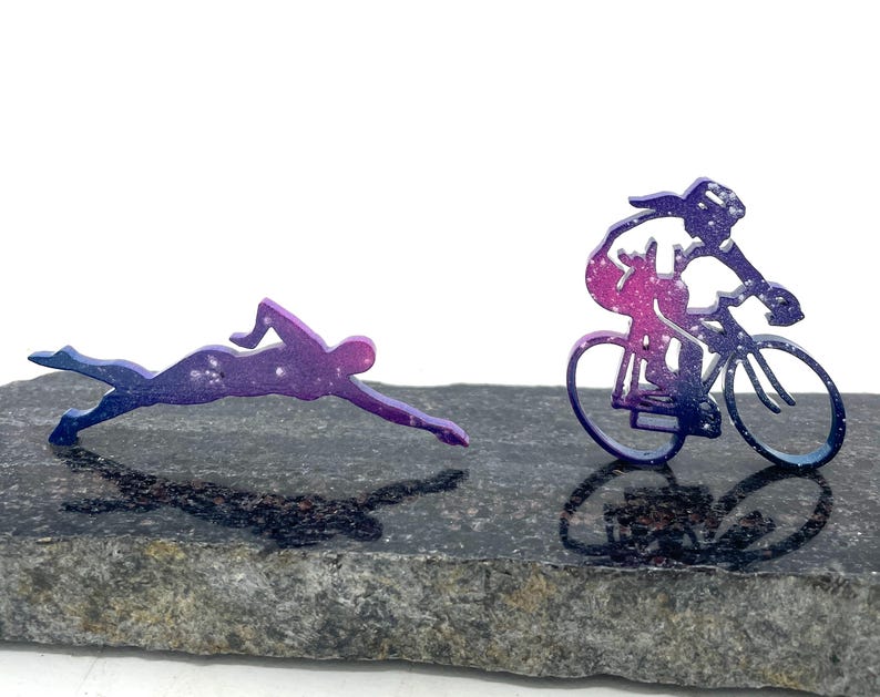 以下が含まれることがあります： 濃い灰色の石の表面に、水泳選手と自転車に乗った人の金属製のシルエットが2つあります。シルエットはピンクとパープルのグラデーションになっています。