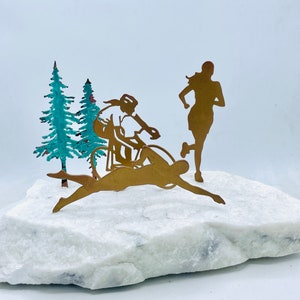 May include: A gold metal sculpture depicting three athletes competing in a triathlon. The sculpture features a swimmer, a cyclist, and a runner. The sculpture is set on a white marble base and is surrounded by two small green trees.