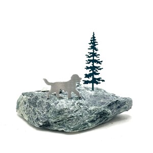 May include: A silver metal dog silhouette standing on a green rock with a dark green metal tree silhouette behind it.