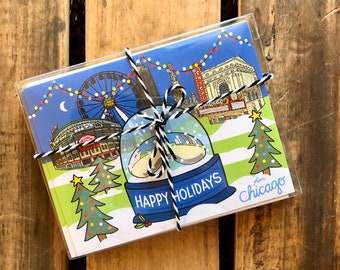 May include: A set of holiday greeting cards featuring a snow globe with a cityscape of Chicago landmarks, including Wrigley Field, the Ferris wheel, and the Chicago Theatre. The cards are tied together with twine and have the text "Happy Holidays from Chicago" printed on them.