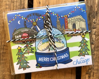 May include: A set of Christmas cards featuring a snow globe with a view of Chicago landmarks, including Wrigley Field, the Ferris wheel, and the Chicago Theatre. The cards are tied together with a white and black twine and have the text "Merry Christmas from Chicago" on the front.