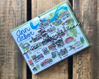 May include: A set of Ann Arbor, Michigan postcards, wrapped in clear cellophane and tied with black and white twine. The postcards feature colorful illustrations of local landmarks and the city's name in blue script.