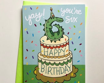 May include: A green alligator wearing a party hat sits on top of a two-tiered cake. The cake is decorated with green, red, and yellow frosting. The card says "yay! you're six happy birthday".