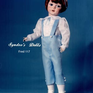 May include: A porcelain doll wearing a blue and white outfit with a blue hat. The doll has brown hair and blue eyes. The doll is labeled "Syndees Dolls Fred 117".