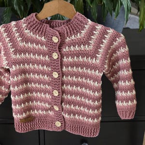 May include: A handmade, button-down, crochet cardigan in shades of mauve and white. The baby sweater features a textured stitch pattern and wooden buttons. The garment is displayed on a wooden hanger.