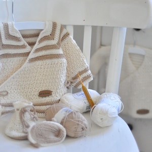 Puede incluir: Un cárdigan de bebé de ganchillo blanco y marrón con botines a juego y ovillos de lana. El cárdigan está atado con una cinta blanca y tiene una etiqueta marrón que dice "Made by Sarah".
