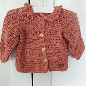 May include: A crocheted cardigan in a dusty rose color with a ruffled collar and button closure. The cardigan has a simple, classic design and is perfect for a baby or toddler.