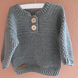 Op de afbeelding: Een grijze gehaakte trui met een ronde hals en lange mouwen. De trui heeft twee houten knopen en een klein, rechthoekig label aan de onderkant. De trui wordt gepresenteerd op een witte hanger.