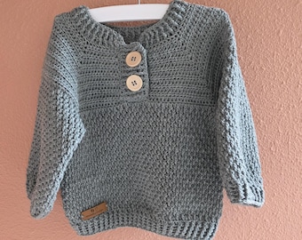 Op de afbeelding: Een grijze gehaakte trui met een ronde hals en lange mouwen. De trui heeft twee houten knopen en een klein, rechthoekig label aan de onderkant. De trui wordt gepresenteerd op een witte hanger.