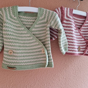 May include: Two crocheted baby cardigans, one green and white striped and one pink and white striped. Both cardigans have a wrap style and button closures.